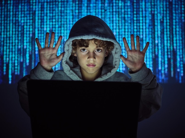 Garoto hacker com as mãos para cima, sentado no laptop no fundo desfocado do código binário na luz escura
