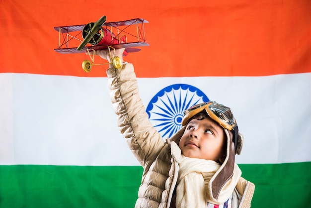 Garoto indiano ou asiático feliz brincando com um avião de metal de brinquedo em trajes e óculos de piloto da 2ª Guerra Mundial, isolado sobre o fundo da bandeira indiana