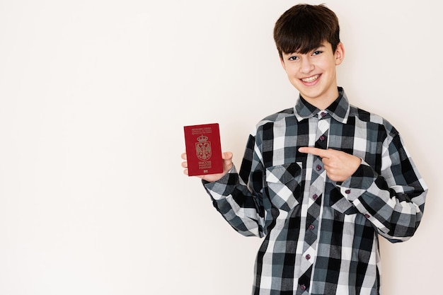 Garoto jovem adolescente segurando o passaporte da Sérvia parecendo positivo e feliz de pé e sorrindo com um sorriso confiante contra o fundo branco