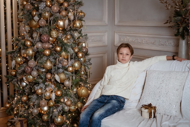 Garoto loiro caucasiano feliz sentado perto da árvore de Natal com um presente