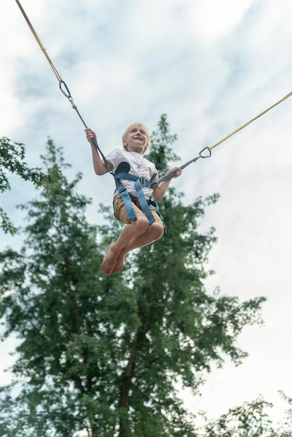 Garoto loiro feliz pendurado em estilingues pula alto no trampolim em um parque de diversões Schoolboy está se divertindo