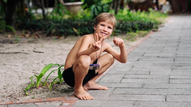 Foto garoto louro sardas agachados sentado sorrindo mostra gesto polegares para cima sem camisa calções de peito nu natureza