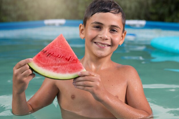 Foto garoto molhado na piscina com uma fatia de melancia vermelha, rindo do conceito de verão