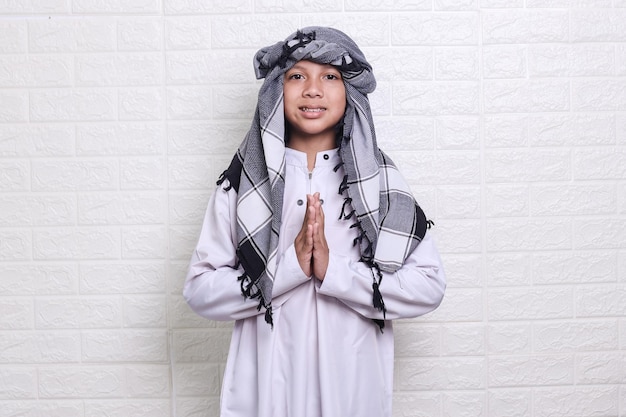 Garoto muçulmano sorridente vestindo túnica branca e calota craniana está de pé e gesticulando para se desculpar com as mãos