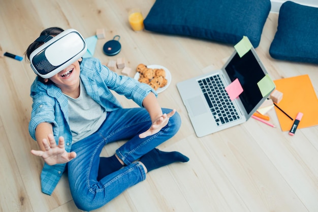 Garoto positivo sentado no chão com um laptop e levantando as mãos enquanto usava óculos de realidade virtual