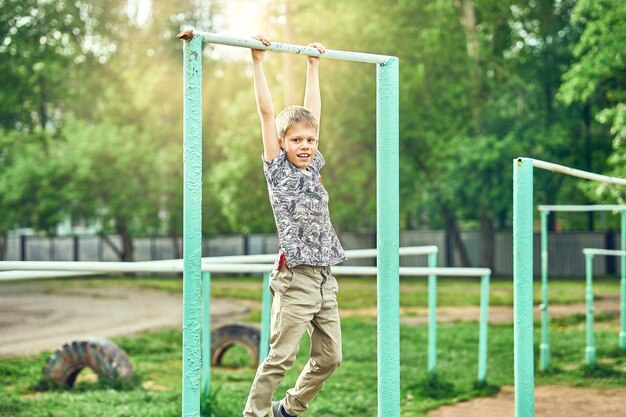 Garoto pré-adolescente forte em roupas casuais fazendo exercícios na barra de metal no campo de esportes no parque