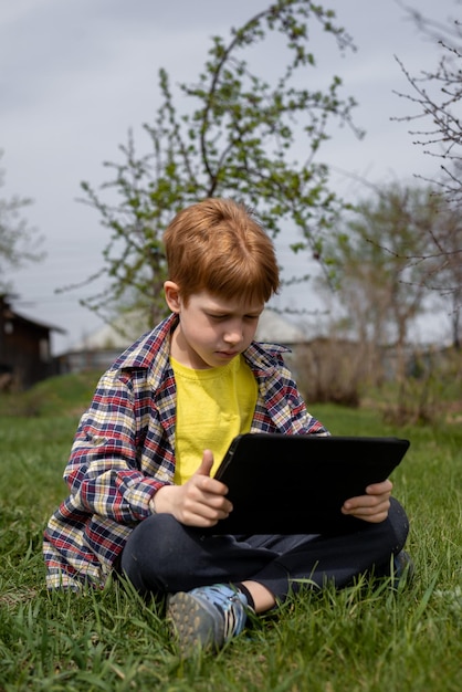 Garoto ruivo feliz jogando tablet ou assistindo desenhos enquanto está sentado na grama verde no quintal na vila de verão closeup