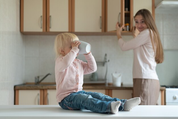 Garoto se senta à mesa da cozinha e bebe da xícara contra o fundo surpreso da mãe.