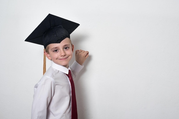 Garoto sorridente bonito usando chapéu de estudante e uniforme escolar criança escreve algo no quadro copiar espaço