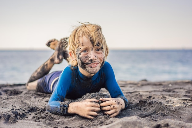 Garoto sorridente do conceito de sexta-feira negra com rosto preto sujo sentado e brincando na praia do mar de areia preta