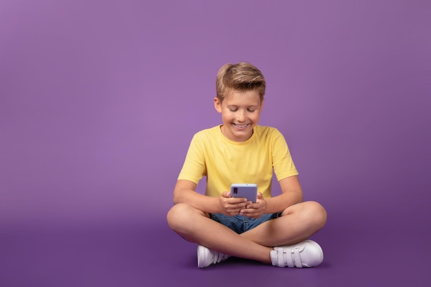 Garoto sorridente pré-adolescente olhando para o celular jogando jogo assistindo vídeo Criança alegre usando celular sentado no chão em fundo roxo Foto de estúdio