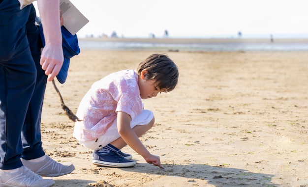 Garoto usando seu dedo escrevendo na areia em um dia quente e ensolarado com pessoas embaçadas andando pelo mar, menino criança brincando na praia do mar com primeiro plano embaçado da perna da avó, férias em família