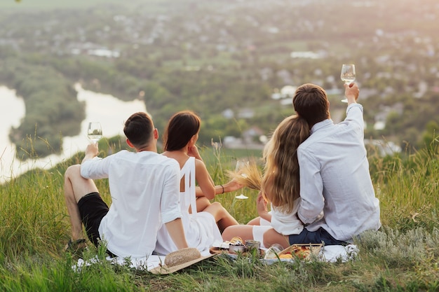 Garotos e garotas em um piquenique romântico, sentados perto de uma cesta de frutas e bebendo vinho branco em taças.