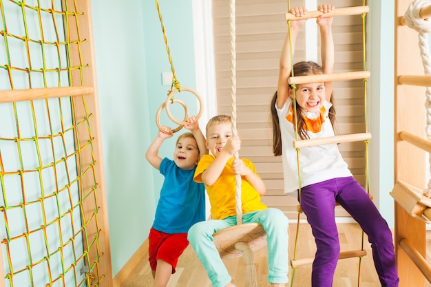 Garotos legais e garota adorável se divertindo, escalando e balançando em uma escada de corda e anéis de madeira em sua sala de treinamento em casa. Adoráveis crianças em roupas coloridas treinando juntos