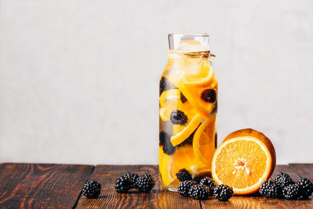 Garrafa de água infundida com laranja crua fatiada e ingredientes frescos de blackberry em mesa de madeira