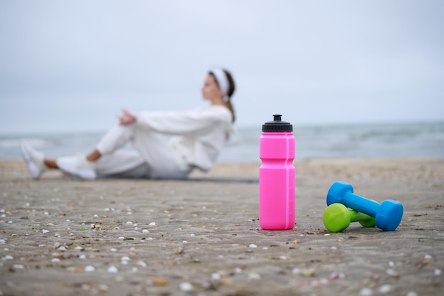 Garrafa de água rosa e halteres na areia Garota fazendo seus exercícios na praia Foto de alta qualidade