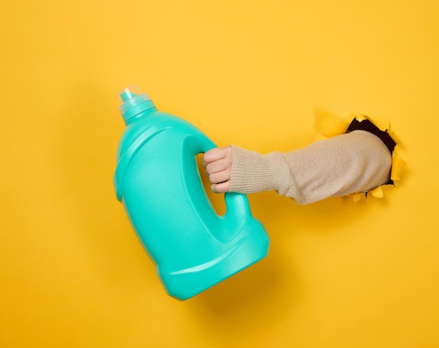Garrafa de plástico azul com detergente líquido em uma mão feminina em um fundo amarelo. Uma parte do corpo sobressai de um buraco rasgado no fundo