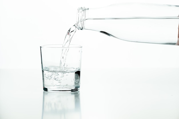 Garrafa de vidro derramando água em um copo meio cheio em um fundo branco
