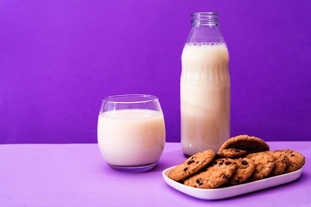 Garrafa de vidro e vidro grande com leite e alguns biscoitos doces de chocolate em um ambiente roxo ou violeta Copiar espaço Orientação horizontal