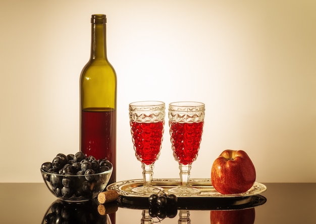 garrafa de vinho tinto duas taças uma maçã e uma taça com uvas em pé sobre a mesa