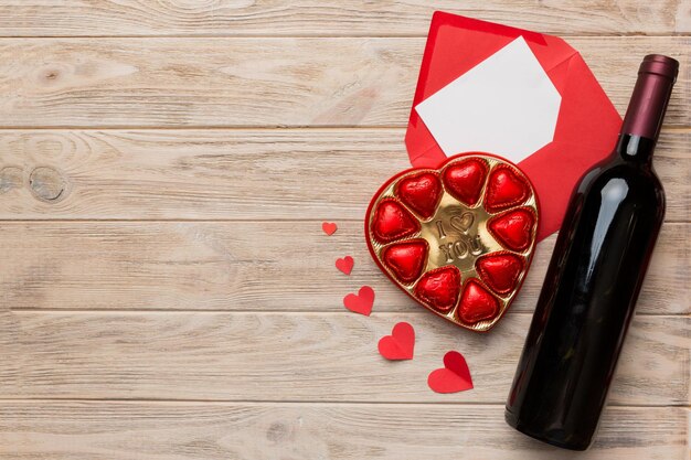 Garrafa de vinho tinto em fundo colorido para o dia dos namorados com envelope de caixa de presente e chocolate em forma de coração com caixa de presente de chocolates vista superior com espaço de cópia
