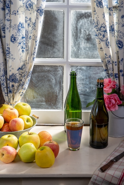Garrafa e copo de cidra com maçãs. perto da janela, na casa rústica
