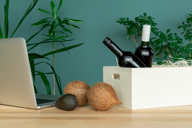 Garrafas de vinho, caixa branca, computador, cocos e abacate na mesa, conceito de pedido online.