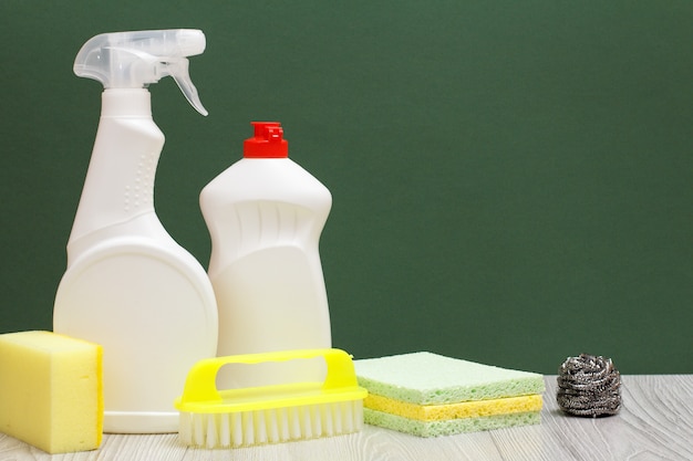 Garrafas plásticas de detergente líquido, limpador de vidros e azulejos, escova, esponjas em fundo verde e de madeira. Conceito de lavagem e limpeza.