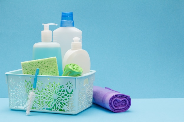Garrafas plásticas de detergente líquido, limpador de vidros e azulejos na cesta, esponjas e sacos de lixo sobre fundo azul. Conjunto de lavagem e limpeza.