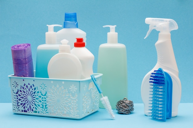 Garrafas plásticas de detergente para louça, limpador de vidros e azulejos na cesta, esponjas, sacos de lixo e escovas sobre fundo azul. Conceito de lavagem e limpeza.