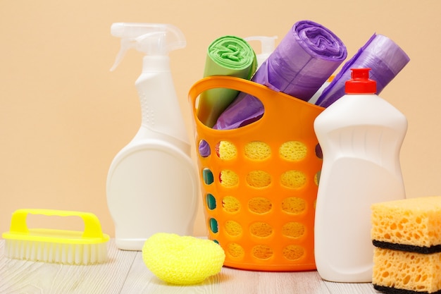 Garrafas plásticas de limpador de vidro e azulejo, cesta com sacos de lixo, esponjas, escova no fundo bege. Conceito de lavagem e limpeza.