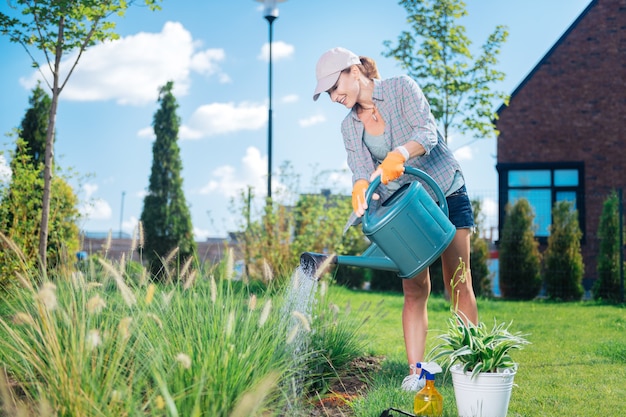 Gartenbett gießen. Blonde Frau mit Schirmmütze und bequemer Freizeitkleidung, die ihr Gartenbett wässert