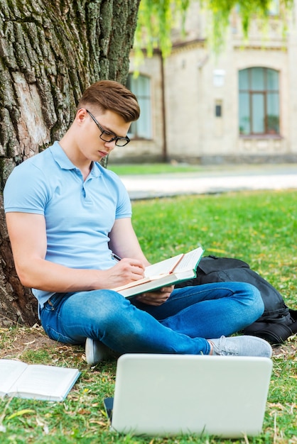 Gastando cada minuto estudando. Estudante do sexo masculino confiante usando óculos escrevendo algo em seu bloco de notas enquanto está sentado na grama e em frente ao prédio da universidade