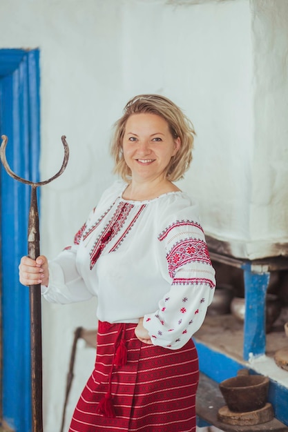 Gastgeberin in bestickter Kleidung mit Schürhaken in der Nähe des Ofens. Retro-Foto des alten ukrainischen Lebens
