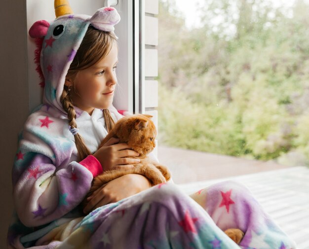 Gatito de jengibre y una niña están sentados en el alféizar de la ventana y mirando por la ventana.