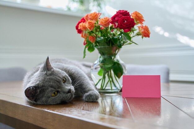 Gato feliz relajado tendido en la mesa con un ramo de flores en un jarrón y una tarjeta con espacio de copia Holiday