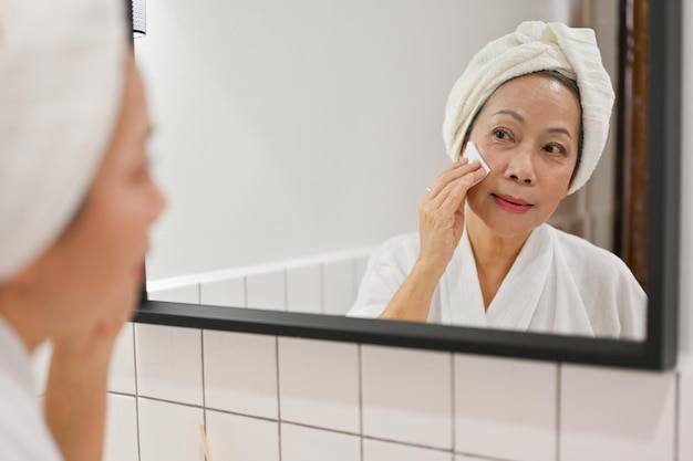 gealterte asiatische Dame, die in den Spiegel schaut und Anti-Aging-Hautpflegebehandlungen mit Wattepad anwendet