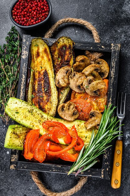 Gebackene Gemüsepaprika, Zucchini, Aubergine und Tomate in einem Holztablett. Schwarzer hölzerner Hintergrund. Ansicht von oben.