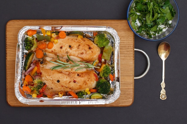 Gebackene Hähnchenbrust oder Filet mit Gemüse und Gemüse in Metallbehälter auf einem Holzbrett. Glasschüssel mit Soße und Metalllöffel. Ansicht von oben.