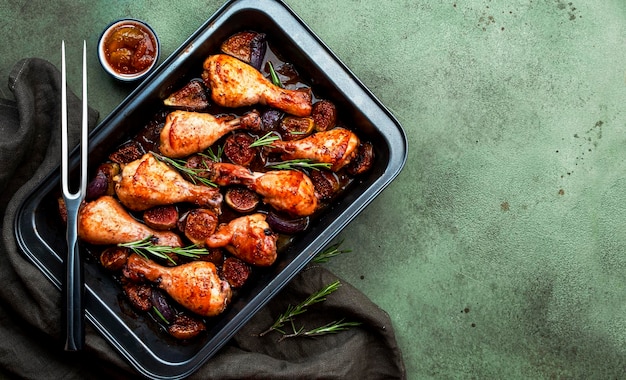 Gebackene Hähnchenkeulen mit süßen Feigen und Zwiebeln in Balsamico-Marinade auf schwarzem Backblech, rostiger grüner Tischhintergrund, Draufsicht