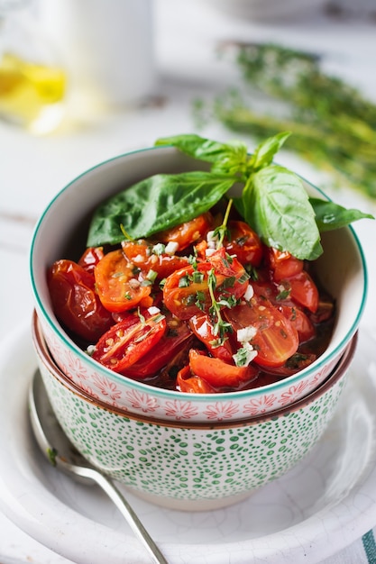 Gebackene Kirschtomate mit Knoblauch, Thymian und Basilikum in Vintage-Keramikschalen auf einem hellen Holztisch. Draufsicht.