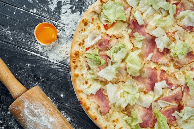 Gebackene Pizza mit Salat, Croutons, weißer Sauce und Hühnchen. Pizza Caesar auf einem schwarzen Holztisch in einer Komposition mit Zutaten. Draufsicht