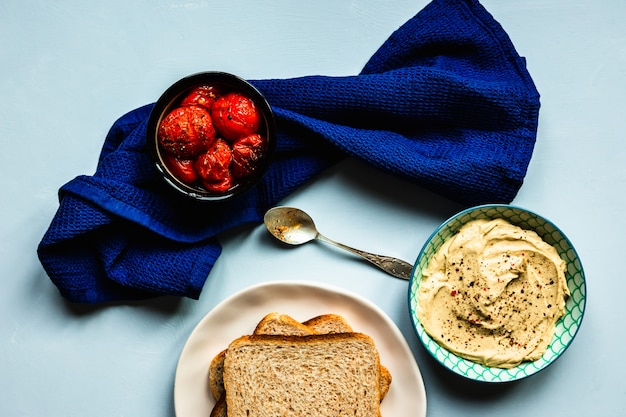 Gebackene Tomaten, Hummus und Brot. Ein tiefblaues Handtuch und ein Löffel. Blaue Oberfläche. Horizontales Bild der Draufsicht.