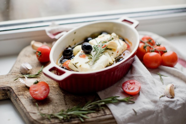 Gebackener Feta-Käse mit Tomaten, Oliven, Knoblauch, Honig und Rosmarin in einer Auflaufform auf rustikalem Hintergrund auf einer Fensterbank