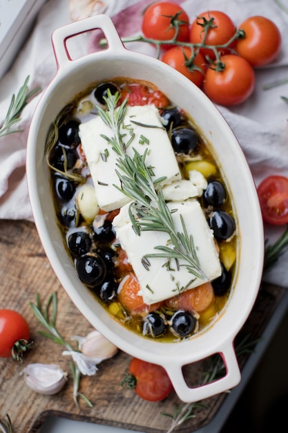 Gebackener Feta-Käse mit Tomaten-Oliven-Knoblauch-Öl-Honig und Rosmarin in Auflaufform auf rustikalem Hintergrund