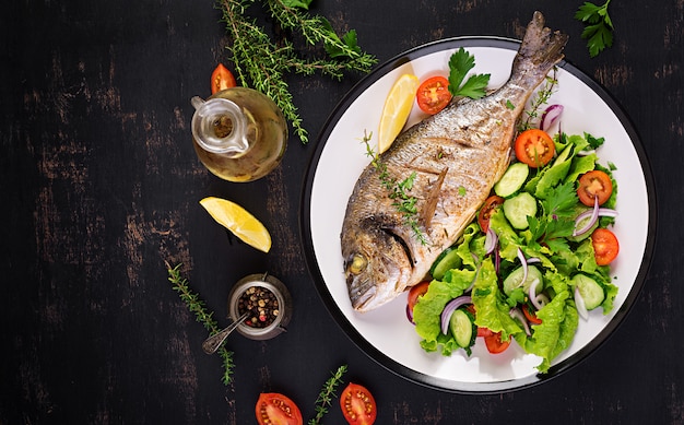 Gebackener Fisch Dorado mit Zitrone und frischem Salat in der weißen Platte auf dunklem rustikalem Hintergrund. Ansicht von oben. Gesundes Abendessen mit Fischkonzept. Abnehmen und sauber essen