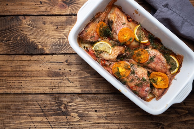 Gebackener Wolfsbarsch mit Tomaten-Pfeffer-Zitrone und Kräutern in einem Keramik-Backblech auf einem Holz