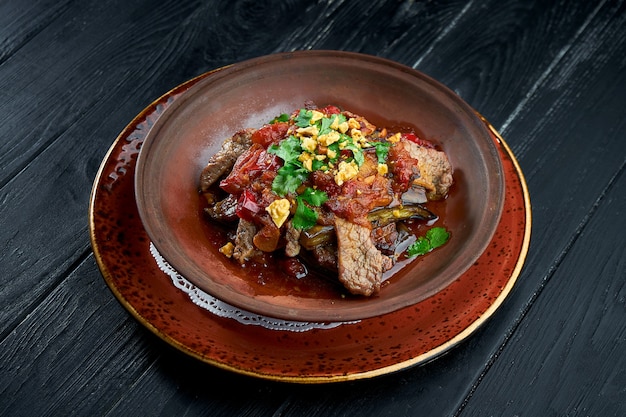 Gebackenes Rindfleisch mit Auberginen, Nüssen, Tomaten und Koriander in Ton, serviert in einer Schüssel auf schwarzem Grund.