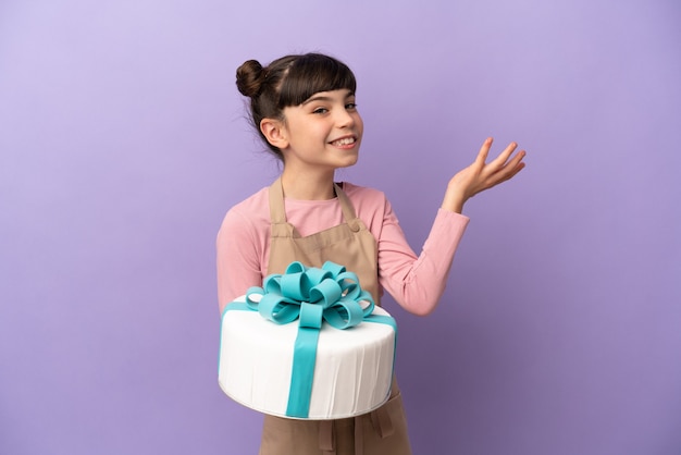 Gebäck kleines Mädchen, das einen großen Kuchen hält, der auf lila Wand lokalisiert wird, die Hände zur Seite ausdehnt, um einzuladen, zu kommen