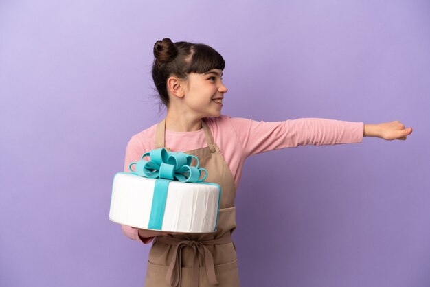 Gebäck kleines Mädchen, das einen großen Kuchen lokalisiert auf lila Wand hält, die eine Daumen hoch Geste gibt